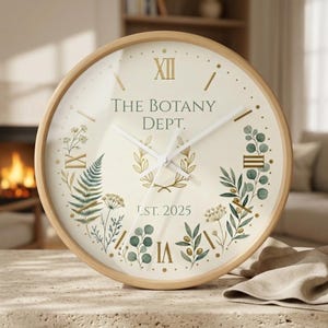 Op de afbeelding: Een ronde wandklok met een licht houten frame. De wijzerplaat is crèmekleurig met gouden Romeinse cijfers en wijzers. De tekst "THE BOTANY DEPT." en "LST. 2025" zijn op de wijzerplaat gedrukt. Botanische illustraties omringen de wijzerplaat.