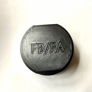 May include: A black, circular object with the letters "FB/FA" embossed on the top. The object has a textured surface with diagonal lines. The item is set against a white background.