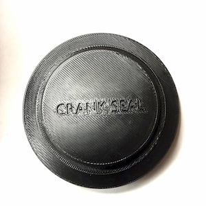 Op de afbeelding: Een zwart, cirkelvormig onderdeel met de tekst "CRANK SEAL" in reliëf. Het onderdeel heeft een geribbelde textuur en een verhoogde buitenrand. Het is waarschijnlijk een mechanisch onderdeel of afdichting.