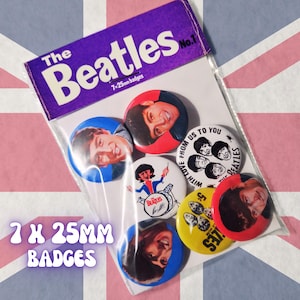 May include: A set of seven 25mm pinback buttons featuring The Beatles, packaged in a clear plastic sleeve with a purple header card labeled The Beatles No. 1. The buttons display individual band member portraits, a cartoon drummer, and group graphics.