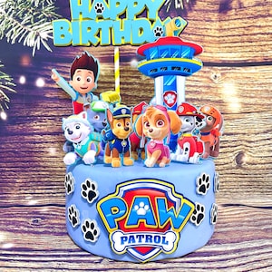 May include: A light blue cake decorated with Paw Patrol characters and the words "Happy Birthday". The cake features the Paw Patrol logo and paw prints. A colorful cake topper with the Paw Patrol theme.
