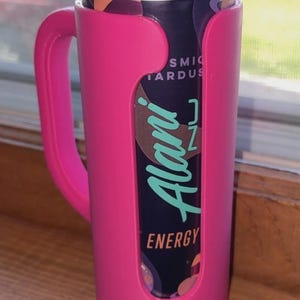 K&ouml;nnte beinhalten: Ein pinkfarbener Getr&auml;nkehalter mit Griff h&auml;lt eine Dose Alani Nu Energy Drink. Die Dose ist lila und schwarz mit dem Alani Nu-Logo in T&uuml;rkis. Der Halter besteht aus Kunststoff und hat einen Griff.