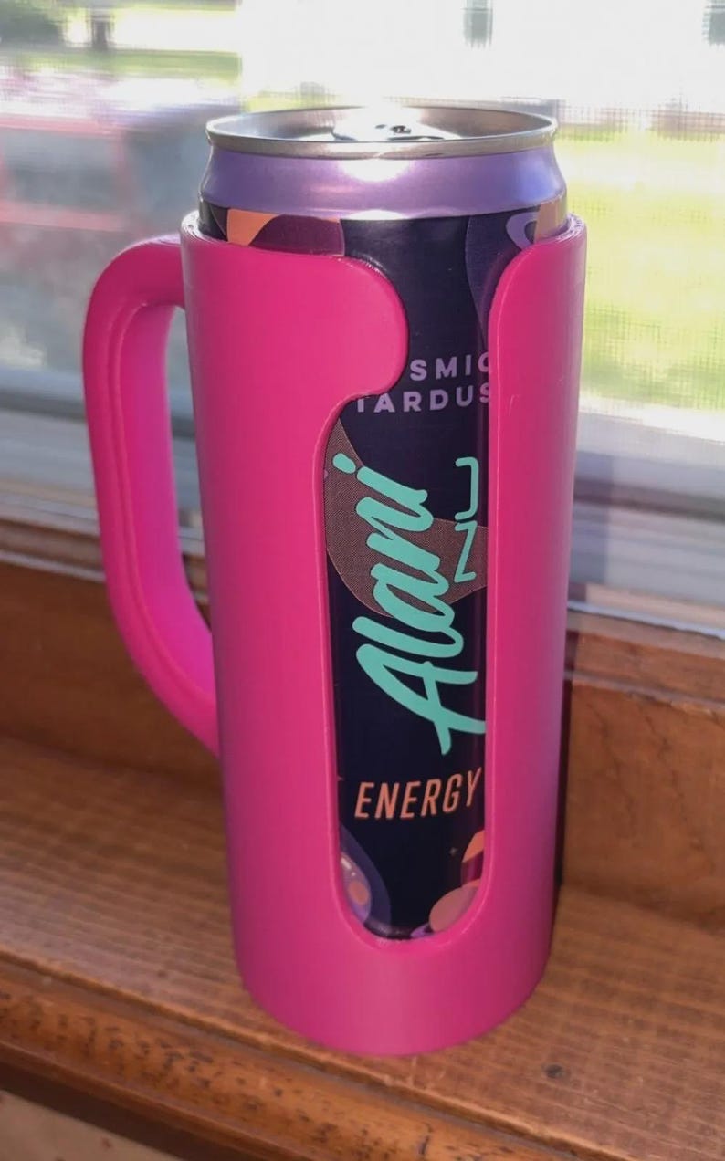 K&ouml;nnte beinhalten: Ein pinkfarbener Getr&auml;nkehalter mit Griff h&auml;lt eine Dose Alani Nu Energy Drink. Die Dose ist lila und schwarz mit dem Alani Nu-Logo in T&uuml;rkis. Der Halter besteht aus Kunststoff und hat einen Griff.