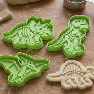 Peut inclure: Trois emporte-pièces en plastique vert vif en forme de squelettes de dinosaures, représentant un tricératops, un tyrannosaure rex et un ptérodactyle, accompagnés d'une pâte à biscuit beige clair en forme de squelette de dinosaure.