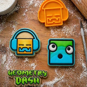 以下が含まれることがあります： Geometry Dashのロゴの形をした3つのクッキー型とクッキー。オレンジ色のプラスチック製の型は、ヘッドホンをつけた四角いキャラクターです。青と黄色のクッキーはヘッドホンをつけた四角いキャラクターで、青い四角いクッキーには目があります。