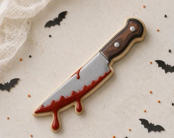 May include: A knife-shaped biscuit decorated with icing to resemble a silver blade with red blood-like drips and a brown wooden handle with two silver rivets. The biscuit is surrounded by small black bat-shaped sprinkles and orange sprinkles.