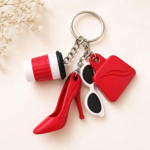 May include: A silver keychain with multiple charms. Charms include a red high heel, a red coffee cup, a pair of white sunglasses with black lenses, and a red lip-shaped charm. The keychain is a fashion accessory.