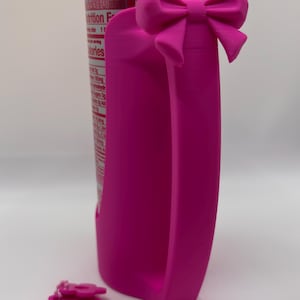 May include: A bright pink plastic can handle with a decorative bow at the top, attached to a beverage can. Beside it lies a matching pink plastic accessory on a small ball chain, featuring a rectangular base and a flower-shaped top detail.