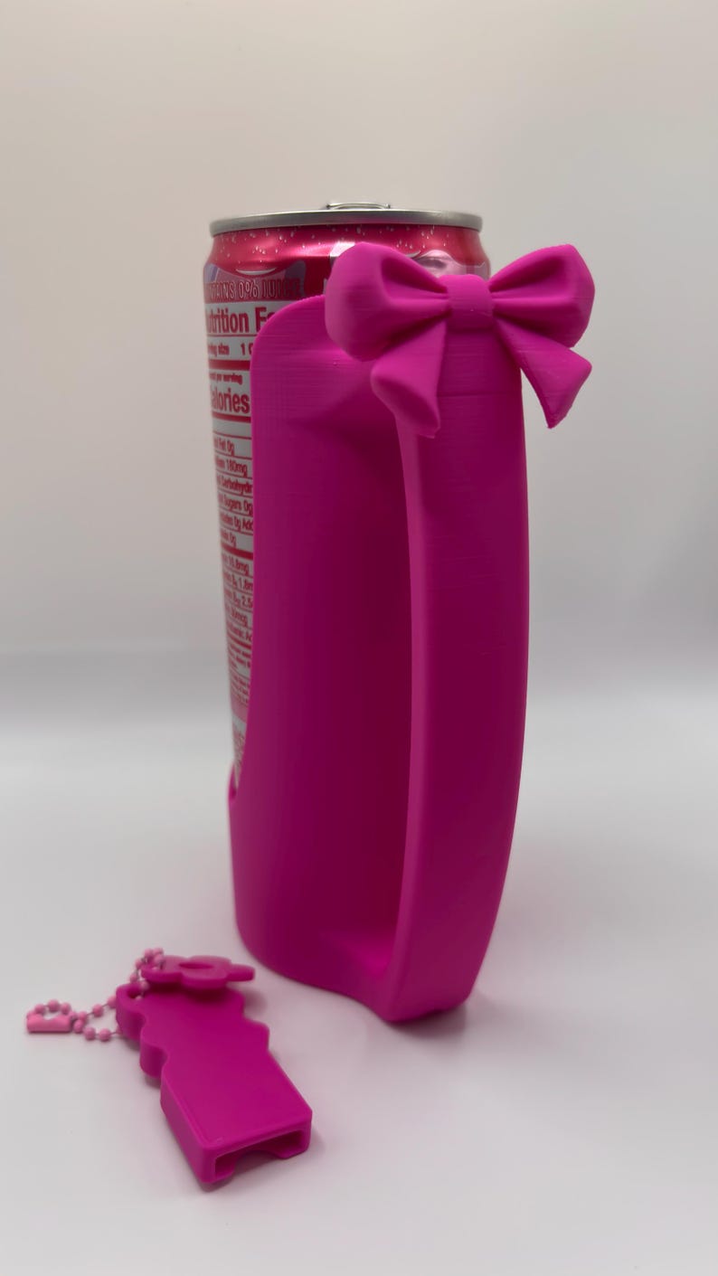 May include: A bright pink plastic can handle with a decorative bow at the top, attached to a beverage can. Beside it lies a matching pink plastic accessory on a small ball chain, featuring a rectangular base and a flower-shaped top detail.