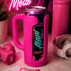 May include: A vibrant pink can cooler with a handle, holding a can of Alani Nu Energy drink. The image includes a pink dumbbell, water bottle, and headphones, with a neon sign that says "Beast Mode". A notebook with the word "FOCUS" is also visible.