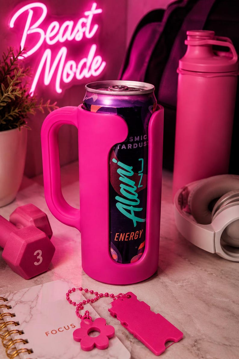 K&ouml;nnte beinhalten: Ein leuchtend rosa Getr&auml;nkek&uuml;hler mit Griff, der eine Dose Alani Nu Energy-Drink h&auml;lt. Das Bild enth&auml;lt eine rosa Hantel, eine Wasserflasche und Kopfh&ouml;rer, mit einem Neon-Schild mit der Aufschrift "Beast Mode". Ein Notizbuch mit dem Wort "FOCUS" ist ebenfalls sichtbar.
