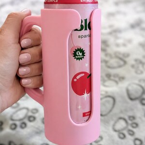 K&ouml;nnte beinhalten: Ein rosa Getr&auml;nkedosenk&uuml;hler mit Griff h&auml;lt eine Dose Shirley Temple-Limonade. Die Dose zeigt eine Kirschgrafik und die Worte "0g Sugar". Der K&uuml;hler besteht aus einem weichen Material.