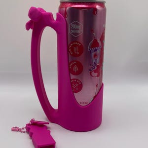 May include: A bright pink silicone can holder with an integrated handle and a decorative bow, holding a pink aluminum beverage can. The can features text including 200mg CAFFEINE, GLUTEN-FREE, VEGAN, and B6 & B12. A small matching pink silicone accessory is placed nearby.