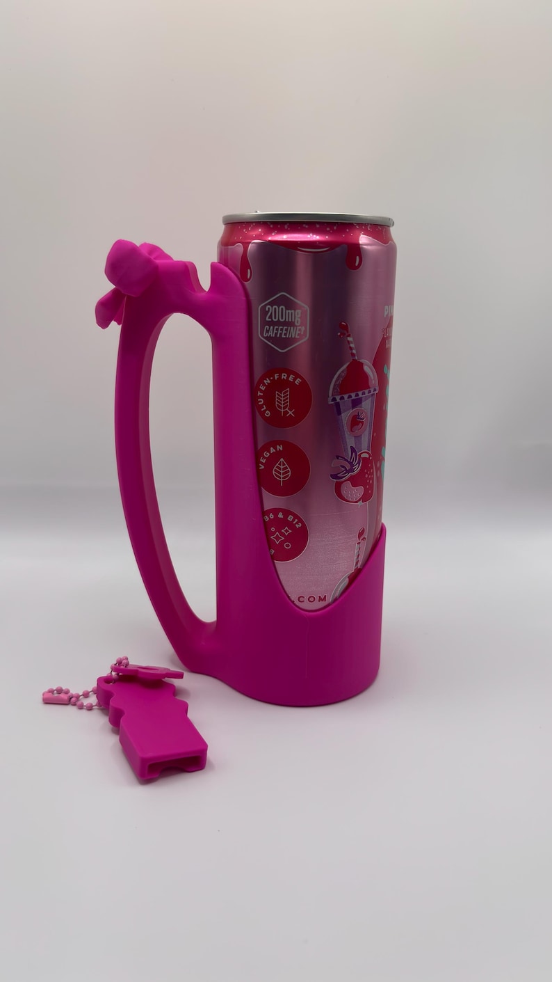 May include: A bright pink silicone can holder with an integrated handle and a decorative bow, holding a pink aluminum beverage can. The can features text including 200mg CAFFEINE, GLUTEN-FREE, VEGAN, and B6 & B12. A small matching pink silicone accessory is placed nearby.