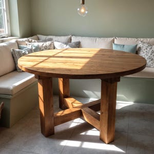 May include: A round wooden dining table with a natural finish. The table has a sturdy base with four thick legs and an X-shaped support. The table is approximately 48 inches in diameter and is placed in a well-lit dining area.
