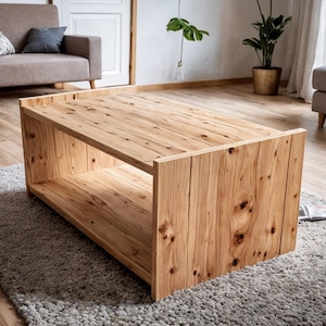 Könnte beinhalten: Ein rechteckiger Couchtisch aus Holz mit einem unteren Regal. Der Tisch besteht aus hellem Holz mit sichtbaren Knoten und Maserungen. Er steht auf einem grauen Teppich in einem Wohnzimmer, mit einem Sofa und Stühlen im Hintergrund.