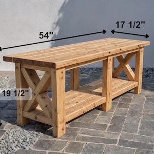DIY Farmhouse X-Base Bench made of light-toned rustic wood featuring a 54-inch length, 17.5-inch depth, and 19.5-inch height. The bench has a thick timber top, decorative X-frame legs, and a lower storage shelf, pictured outdoors on a stone patio."