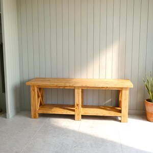May include: A light-colored wooden bench with a lower shelf and X-shaped supports. The bench is in front of a light gray wall. A potted plant and a closet are also visible.