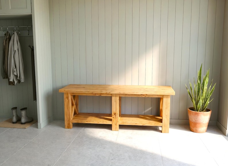 May include: A light-colored wooden bench with a lower shelf and X-shaped supports. The bench is in front of a light gray wall. A potted plant and a closet are also visible.