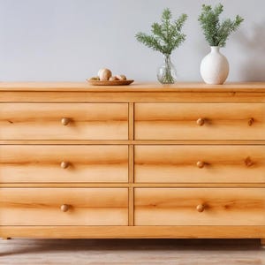Könnte beinhalten: Eine hellfarbene Holzkommode mit sechs Schubladen und runden Holzknöpfen. Die Kommode ist mit einem kleinen Holztablett mit mehreren runden Gegenständen belegt. Zwei Vasen mit Grünzeug stehen oben.