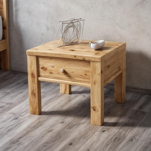 Puede incluir: Una mesita de noche de madera clara con un solo cajón y un pequeño pomo redondo. Una escultura decorativa de metal y un pequeño cuenco blanco encima. La mesita forma parte de un juego de muebles de dormitorio.