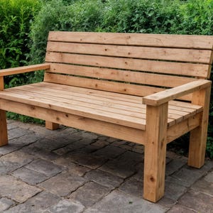 Könnte beinhalten: Eine Gartenbank aus Holz mit natürlicher Oberfläche. Die Bank hat eine Sitzfläche und Rückenlehne mit Lamellen sowie Armlehnen. Die Bank ist aus hellfarbigem Holz gefertigt und steht auf einer Steinpflasterung mit grünem Laub im Hintergrund.