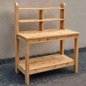 May include: A wooden potting bench with a natural finish. It features a tabletop, a lower shelf, and a back with two shelves. The bench is constructed from light-colored wood and is designed for outdoor use.