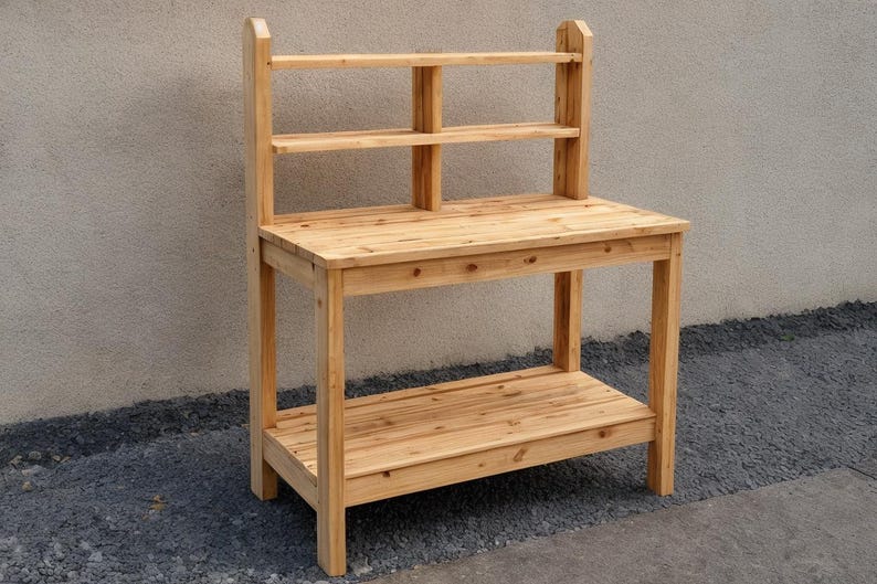 May include: A wooden potting bench with a natural finish. It features a tabletop, a lower shelf, and a back with two shelves. The bench is constructed from light-colored wood and is designed for outdoor use.