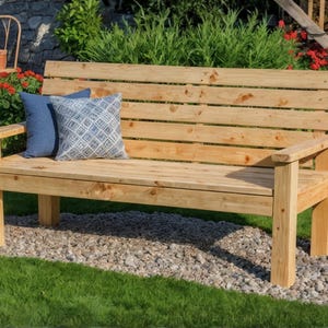May include: A wooden garden bench with a slatted back and armrests. Two blue cushions with geometric patterns are placed on the bench. The bench is made of light-coloured wood and sits on a bed of small stones.