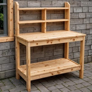 May include: A light-colored wooden potting bench with a shelf and two upper shelves. The bench has a rectangular tabletop and a lower shelf for storage. The background is a gray wooden wall and a brick patio.