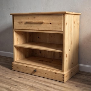 May include: A light-colored wooden cabinet with a drawer and two shelves. The cabinet is made of pine and has a natural finish. The drawer has a simple, rectangular handle. The cabinet is designed for storage and display.