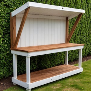May include: A wooden potting bench with a white frame and a brown tabletop and shelf. The bench has a white slatted back and a white angled roof supported by brown wooden beams. The bench is outdoors on a grassy lawn.