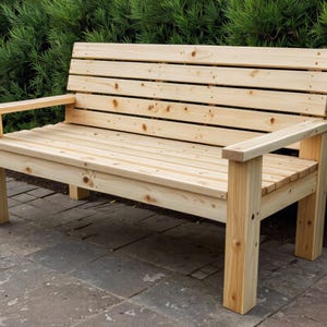 May include: A wooden outdoor bench with a natural finish. The bench has a slatted seat and backrest, with armrests. The bench is made of light-colored wood and is set against a backdrop of green foliage.