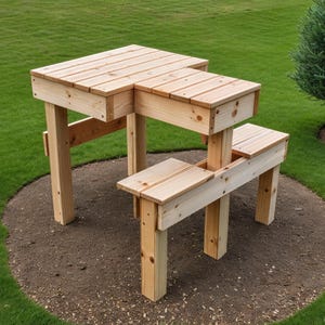 May include: A light-colored wooden picnic table with an attached bench. The table has a rectangular top with a step-down design. The bench is connected to the table's side. The table and bench are made of light-colored wood and are set on a circular bed of mulch.