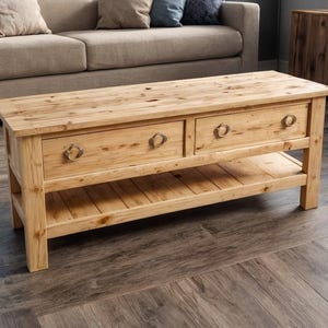 May include: A light-colored wooden coffee table with two drawers and a lower shelf. The table has a rectangular shape and is supported by four sturdy legs. The drawers have ring-pull handles. The table measures approximately 48 inches long, 24 inches wide, and 18 inches high.