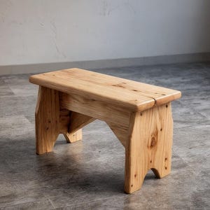 May include: A small, handcrafted wooden bench with a natural finish. The bench has a rectangular seat and angled legs, showcasing the wood grain. It is approximately 61 cm long, 25 cm wide, and 30 cm high.