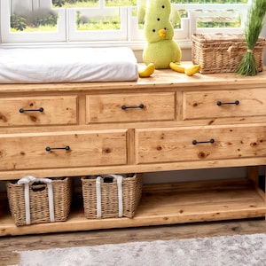 Puede incluir: Una cómoda de madera de color claro con siete cajones y dos cestas de mimbre. La cómoda tiene un cambiador blanco en la parte superior y herrajes negros. Un peluche y objetos decorativos están en la parte superior.