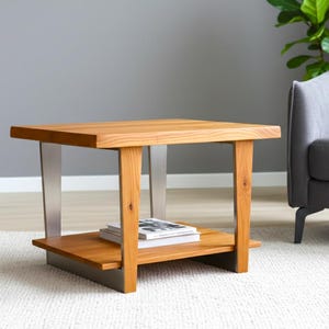 May include: A modern, square coffee table with a natural wood finish. The table has a lower shelf and is supported by metal and wood legs. A stack of books rests on the lower shelf. The table is approximately 30 inches long.