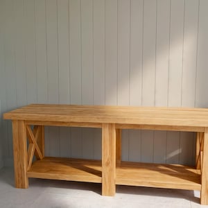 May include: A light-colored wooden bench with a lower shelf and X-shaped supports on the sides. The bench is made of solid wood and has a natural finish. The background is a light gray wall.