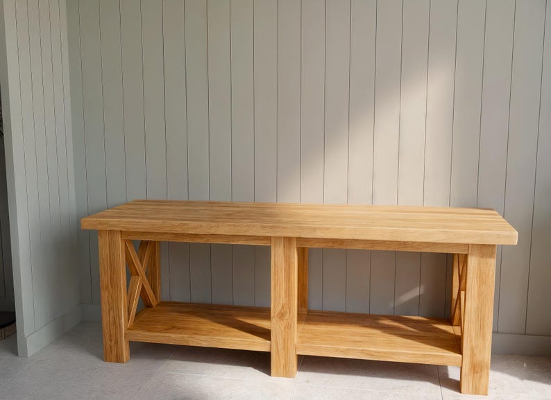 May include: A light-colored wooden bench with a lower shelf and X-shaped supports on the sides. The bench is made of solid wood and has a natural finish. The background is a light gray wall.