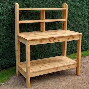 May include: A wooden potting bench with a natural finish. The bench has a tabletop, a lower shelf, and a back with two small shelves. The structure is made of light-colored wood and is designed for outdoor use.