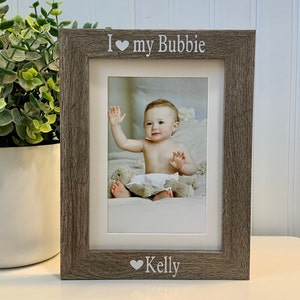 May include: A rustic wood picture frame with a white mat and a photo of a baby. The frame has the text "I love my Bubbie" and "Kelly" with a heart symbol.