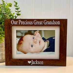 May include: A brown wooden picture frame with a white mat and a photo of a baby boy. The frame has the text "Our Precious Great Grandson" at the top and "Jackson" with a heart symbol at the bottom.