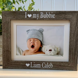May include: A rustic wood picture frame with a white background. The frame has a white inscription that reads "I ♥ my Bubbie" and "♥ Liam Caleb". The frame contains a photo of a baby wearing a blue hat and yawning, with a white teddy bear beside them.