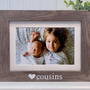 May include: A rustic brown wood picture frame with a white mat and a photo of two children, a baby and a young girl, lying on a patterned blanket. The frame has a white heart and the word "cousins" printed on the bottom.