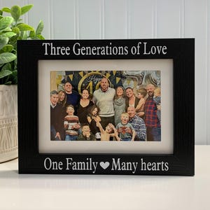 May include: A rectangular black picture frame with white text engraved on the top and bottom borders. The top border reads Three Generations of Love and the bottom border reads One Family Heart Many hearts. The frame holds a color photograph of a large family.