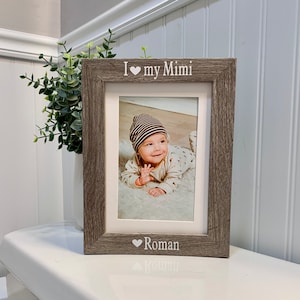 May include: A rectangular picture frame with a weathered gray wood finish. The top border is engraved with the text I heart my Mimi, and the bottom border features a heart symbol followed by the name Roman. The frame holds a photo of a baby.
