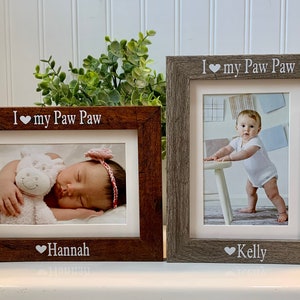 May include: Two wooden picture frames with the text "I love my Paw Paw" at the top. The left frame has a photo of a baby girl sleeping with a stuffed animal and the name "Hannah" at the bottom. The right frame has a photo of a baby girl standing and the name "Kelly" at the bottom.