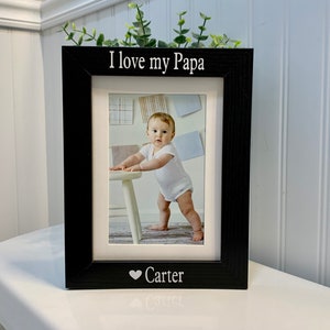 May include: Black picture frame with a white mat and a photo of a baby standing in front of a white chair. The frame has the text "I love my Papa" at the top and "Carter" with a heart symbol at the bottom.