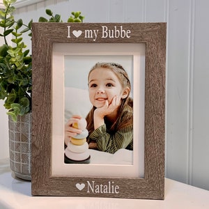 May include: A wood-toned picture frame with a white mat and a photo of a young girl. The frame has the text "I love my Bubbe" and "Natalie" with a heart symbol.